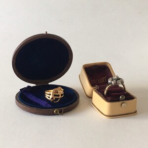 Op de afbeelding: Twee ringendozen, een bruine met een gouden ring erin en een beige met een diamanten ring erin. De beige doos heeft de tekst "Tiffany & Co." erop.