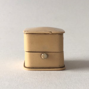 Op de afbeelding: Een klein, vierkant, cr&egrave;mekleurig doosje met een scharnierend deksel en een kleine, ronde knop aan de voorkant.