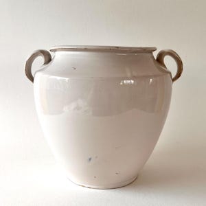 Op de afbeelding: Een grote, gebroken witte keramische vaas met twee gebogen handvatten. De vaas heeft een brede body die taps toeloopt naar de basis en een licht uitlopende rand. De handvatten en rand zijn een donkerdere tint beige. Het oppervlak heeft een glanzende afwerking.