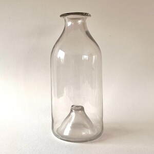May include: A clear glass bottle with a unique design. The bottle has a smaller, inverted bottle shape at the bottom.