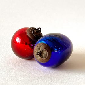 Op de afbeelding: Twee ronde glazen ornamenten, een rode en een blauwe, elk voorzien van een verweerde, donker metalen dop met een klein draadlusje om op te hangen. De oppervlakken van de ornamenten zijn glad en reflecterend met duidelijke lichtvlekken.
