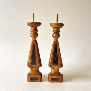 Op de afbeelding: Twee antieke, goudkleurig geschilderde houten kandelaars. Elk heeft een gelaagde basis, een zwart driehoekig accent en een punt aan de bovenkant. De kandelaars worden tegen een witte achtergrond getoond.
