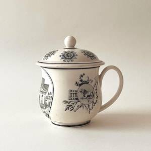 Könnte beinhalten: Cremefarbene Keramiktasse mit Deckel, mit schwarzen Illustrationen eines Gebäudes, eines Löwen und floralen Mustern. Die Tasse hat einen Henkel und einen dekorativen Rand. Der Deckel hat einen kleinen Knopf und ein sich wiederholendes geometrisches Design.
