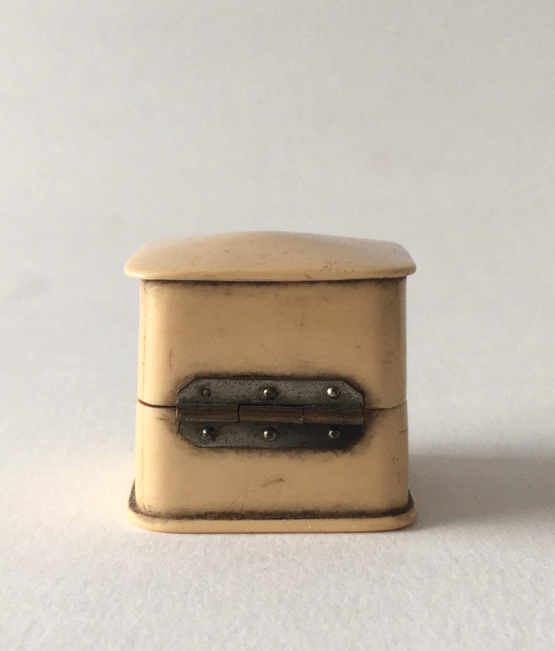 Op de afbeelding: Een klein, vierkant, ivoren doosje met een scharnierend deksel. Het doosje heeft een metalen scharnier met decoratieve klinknagels.