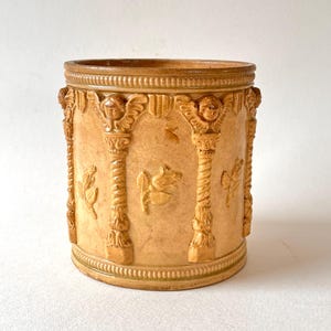 Op de afbeelding: Een cilindervormige, beige keramische pot met een reliëfpatroon van verticale zuilen bekroond met cherubijngezichten. Het buitenoppervlak is versierd met in reliëf aangebrachte bloemmotieven en een kralenrand langs de boven- en onderzijde.