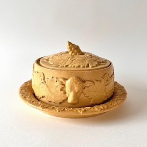 Op de afbeelding: Een ronde, beige keramische botervloot met bijpassend deksel. De buitenkant van de schaal en het deksel zijn voorzien van reliëfdetails van wijnbladeren en een koeienkop. Het deksel is bekroond met een kleine, gebeeldhouwde tros druiven.