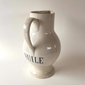 Könnte beinhalten: Cremefarbener Keramikkrug mit rundem Körper, Henkel und Ausguss. Das Wort "HUILE" ist in Schwarz aufgedruckt. Der Krug steht vor weißem Hintergrund.