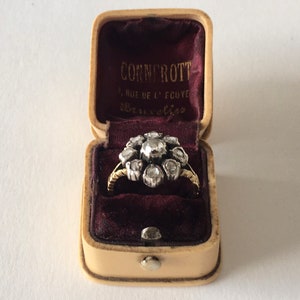 Op de afbeelding: Een vintage diamanten ring in een cr&egrave;mekleurige ringdoos met een bordeauxrode fluwelen binnenkant. De doos is gelabeld "Connrott, 4 Rue de l'Ecuyer, Bruxelles".
