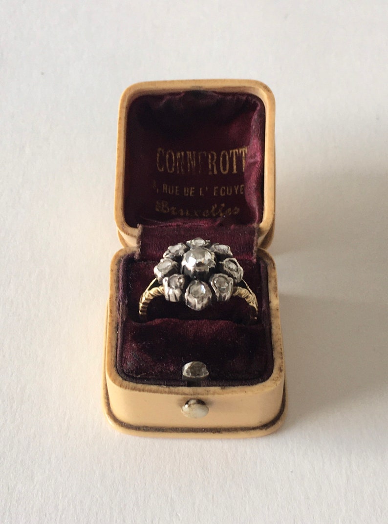 Op de afbeelding: Een vintage diamanten ring in een cr&egrave;mekleurige ringdoos met een bordeauxrode fluwelen binnenkant. De doos is gelabeld "Connrott, 4 Rue de l'Ecuyer, Bruxelles".
