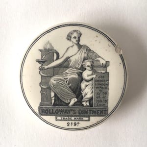 Könnte beinhalten: Ein runder weißer Keramikdeckel mit einem schwarzen Aufdruck einer sitzenden Figur und einem Kind neben einer Säule mit einer Schlange. Der Text lautet HOLLOWAY'S OINTMENT, TRADE MARK, 533 OXFORD ST LONDON und 2s 9d, sowie weitere Preisangaben.