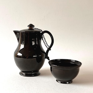 Op de afbeelding: Een glanzende zwarte keramische theepot met een bijpassend deksel dat is vastgezet met een klein metalen kettinkje, voorzien van een gebogen handvat en een tuit. Naast de theepot staat een kleine, bijpassende zwarte keramische kom met ronde voet.