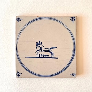 Può includere: Piastrella quadrata in ceramica con un design blu e bianco. La piastrella presenta un contorno blu di un cavallo che salta un recinto all'interno di un bordo circolare blu. Accenti floreali blu decorativi sono in ogni angolo.