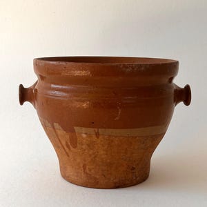 May include: A terracotta pot with two handles. The pot is brown with a light brown glaze. It has a rounded bottom and a wide opening.