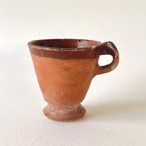 Op de afbeelding: Een kleine, handgemaakte terracotta beker met een handvat. De beker heeft een uitlopende vorm, een donkerbruine rand en een voet. De buitenkant heeft een warme, aardse oranjebruine kleur, met een rustieke, getextureerde afwerking.