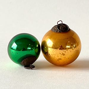 Op de afbeelding: Twee bolvormige glazen ornamenten, één in levendig smaragdgroen en de andere met een reflecterende gouden afwerking. Elk ornament heeft een donkere, verweerde metalen dop aan de bovenkant met een kleine ronde lus om op te hangen.