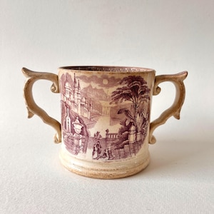 Può includere: Tazza in ceramica color crema con due manici, con una scena di paesaggio bordeaux. La scena raffigura un castello, alberi e figure. Il bordo presenta un motivo a chiave greca.