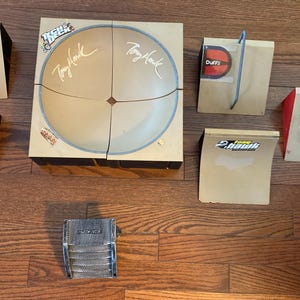 May include: A collection of miniature skateboard ramps and a half-pipe, featuring the "Tony Hawk" logo and signature. The set includes ramps with "Duffs" and "Haro" branding, and a metal skateboard accessory. The items are arranged on a wooden surface.