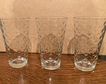 May include: Three clear glass tumblers with a textured, bubbled surface. The glasses are empty and sitting on a wooden table.