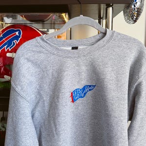May include: A grey sweatshirt with the text "Lets Go Buffalo" in blue and red. The sweatshirt is hanging on a grey hanger. In the background are miniature American football helmets and a disco ball.