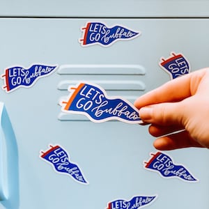 May include: A collection of blue, white, and red pennant-shaped stickers with the text "Lets Go Buffalo" in a script font. The stickers are on a light blue surface, with one being held by a hand.