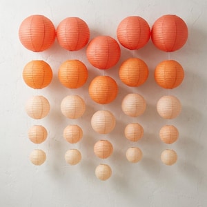 May include: A collection of spherical paper lanterns arranged in five vertical columns. The lanterns feature a ribbed texture and transition in color from deep orange at the top to a pale, creamy peach shade at the bottom of each string.