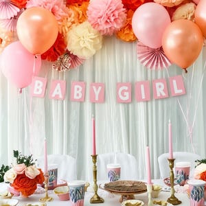 May include: A festive baby shower scene with pink and peach balloons, paper flowers, and a banner that reads "BABY GIRL." The table is set with pink plates, cups, and candles, creating a celebratory atmosphere. Floral arrangements and decorative elements complete the party decor.