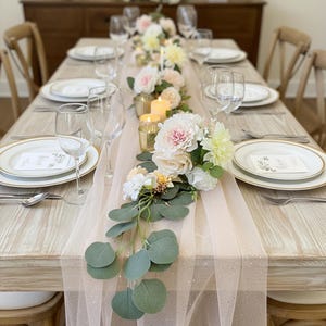 May include: A beautifully set dining table with a light pink tulle table runner. The table is adorned with white plates, wine glasses, silverware, and a floral centerpiece with pink and white flowers, candles, and greenery. The table is set for a special occasion.