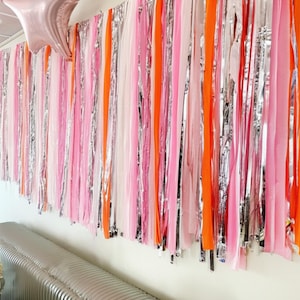 May include: A decorative wall hanging featuring vertical strips of pink, orange, silver, and white fabric. A pink star-shaped balloon is suspended near the top. The wall hanging is likely used for party or event decorations.