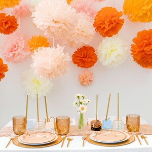 May include: A festive table setting with gold-rimmed plates, cutlery, and textured glasses. The table is decorated with a peach-colored runner and small flower arrangements. Overhead, large paper pom-poms in shades of peach, orange, and white create a celebratory atmosphere.