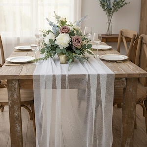 May include: A rustic wooden dining table set for a meal, featuring a white sheer table runner with a subtle glitter pattern. A floral centerpiece with white, mauve, and blue flowers sits atop the runner. White plates and wine glasses complete the setting.