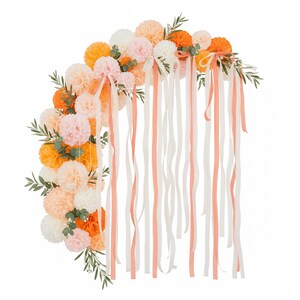 May include: A semi-circular floral arrangement featuring orange, peach, pink, and white pom-poms, accented with green foliage. Long, flowing ribbons in white and peach cascade downwards, creating a decorative and festive display.