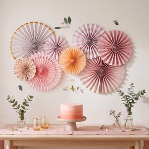 May include: A festive scene featuring a pink ombre cake on a pedestal, surrounded by paper fans in shades of pink, peach, and gold. Eucalyptus and small vases with flowers decorate the table, creating a celebratory atmosphere.