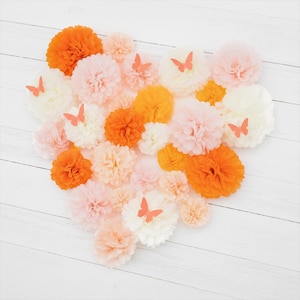 May include: An assortment of colorful paper pom-poms in shades of orange, peach, and white, arranged in a heart shape. Small orange butterfly cutouts are placed on some of the pom-poms. The background is a white wooden surface.