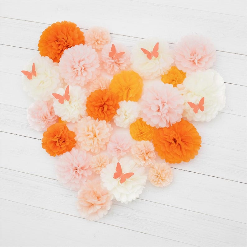 May include: An assortment of colorful paper pom-poms in shades of orange, peach, and white, arranged in a heart shape. Small orange butterfly cutouts are placed on some of the pom-poms. The background is a white wooden surface.
