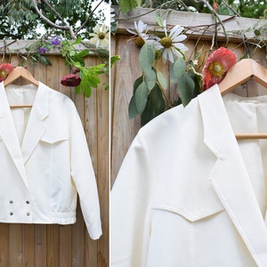 Op de afbeelding: Een wit getailleerd blazer met een dubbele rij knopen en twee klepzakken. De blazer heeft een licht geplooide zoom en hangt aan een houten hanger.