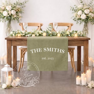 Peut inclure: Une table de réception de mariage en bois, ornée d'un chemin de table vert avec l'inscription "THE SMITHS est. 2023". La table est décorée de fleurs, de bougies et de deux chaises. Des fleurs blanches sont dans des vases de chaque côté.