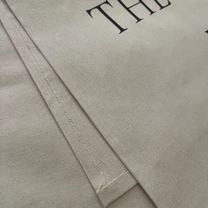 May include: A beige canvas tote bag featuring black text that reads THE O'B. The fabric has a visible woven texture with stitched seams along the edges. The material is a light tan color, and the lettering is printed in a bold, serif font style.
