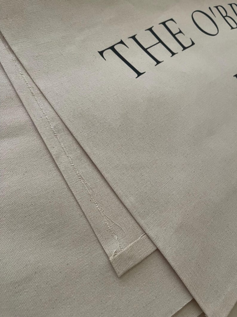 May include: A beige canvas tote bag featuring black text that reads THE O'B. The fabric has a visible woven texture with stitched seams along the edges. The material is a light tan color, and the lettering is printed in a bold, serif font style.