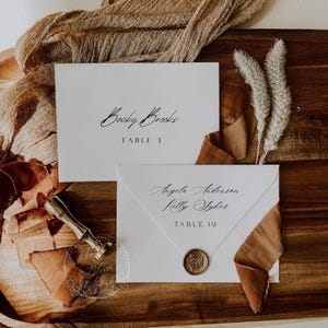 May include: Elegant wedding stationery featuring two white cards with calligraphy names and table numbers. One card reads "Becky Brooks, TABLE 3." The other reads "Angela Anderson, Kelly Sykes, TABLE 19." Includes a wax seal and a decorative ribbon.