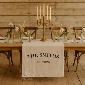 May include: A beige fabric table runner featuring the black printed text THE SMITHS and est. 2026. The runner is displayed on a wooden table with a gold five-arm candelabra, clear glass vases containing pink roses and dried grasses.