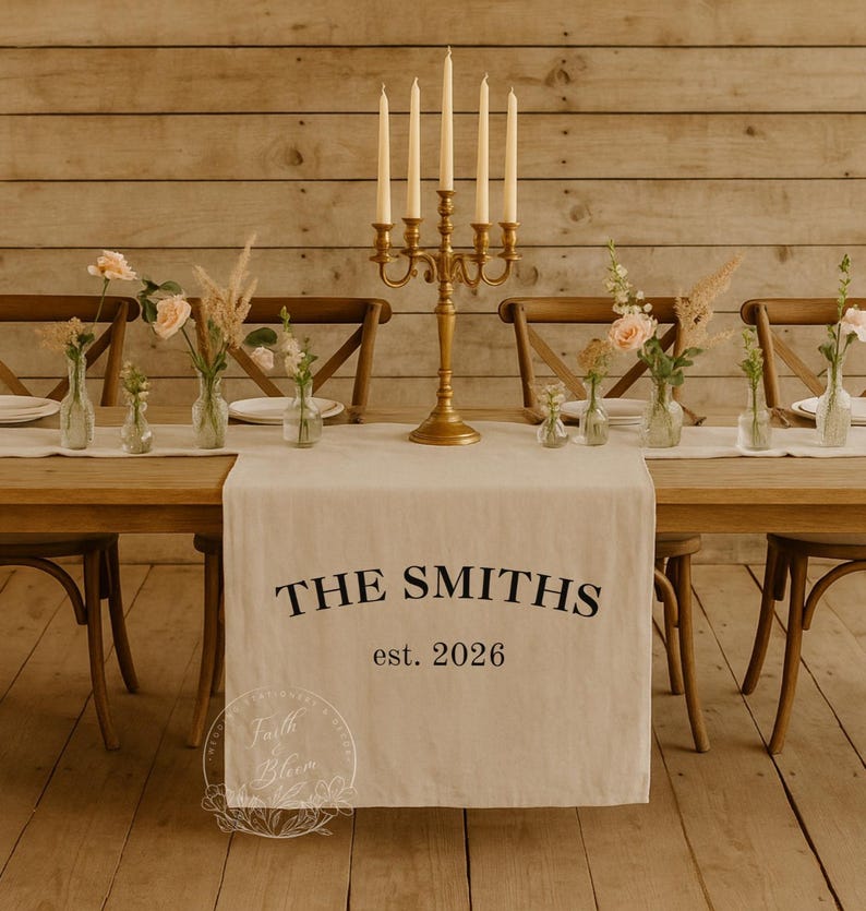May include: A beige fabric table runner featuring the black printed text THE SMITHS and est. 2026. The runner is displayed on a wooden table with a gold five-arm candelabra, clear glass vases containing pink roses and dried grasses.