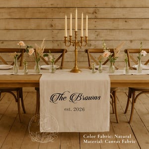 May include: A natural-colored canvas table runner featuring the black printed text The Browns est. 2026. The runner is displayed on a wooden table with a gold five-arm candelabra and several small glass vases containing flowers and dried grasses.