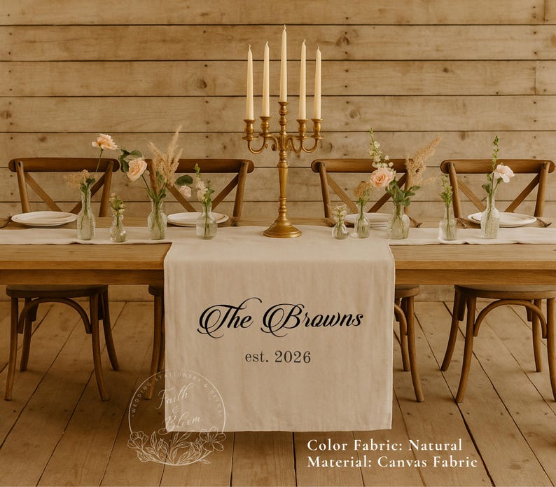 May include: A natural-colored canvas table runner featuring the black printed text The Browns est. 2026. The runner is displayed on a wooden table with a gold five-arm candelabra and several small glass vases containing flowers and dried grasses.