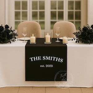 Puede incluir: Una mesa de recepción de bodas está decorada con un mantel blanco y un camino de mesa negro. El camino de mesa muestra "THE SMITHS est. 2023" en texto blanco. Arreglos florales de rosas negras y velas decoran la mesa.