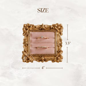 May include: A square gold-colored ring display holder with an ornate, decorative frame and a soft pink velvet interior featuring two horizontal slots. The display measures 4 inches in width and 3.5 inches in height, holding two gold rings.