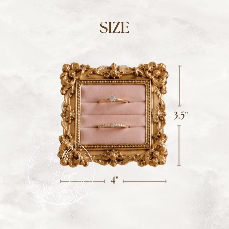 May include: A square gold-colored ring display holder with an ornate, decorative frame and a soft pink velvet interior featuring two horizontal slots. The display measures 4 inches in width and 3.5 inches in height, holding two gold rings.