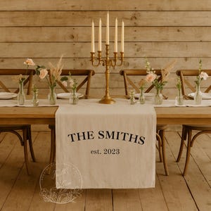 Puede incluir: Un camino de mesa de tela color crema con el texto impreso en negro THE SMITHS y est. 2023. El camino se muestra sobre una mesa de madera con un candelabro dorado de cinco brazos, pequeños jarrones de cristal con flores y platos blancos.