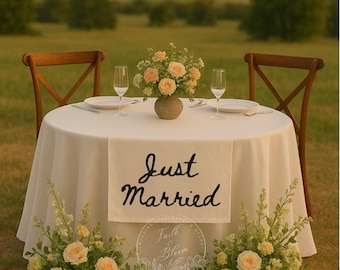 Op de afbeelding: Een ronde tafel met een wit tafelkleed en de tekst Just Married in zwarte schrijfletters. De tafel is gedekt met twee witte borden, twee glazen en een vaas met perzikkleurige rozen. De tafel wordt geflankeerd door twee houten stoelen.