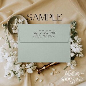 May include: A light blue envelope with a black ink return address stamp that reads "soon to be Mr. & Mrs. Hill 726 Jack Ave Tampa, FL 33592". The envelope is lying on a beige surface with white flowers.