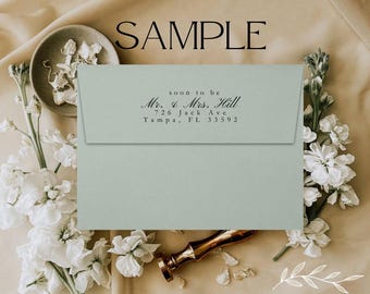 May include: A light blue envelope with a black ink return address stamp that reads "soon to be Mr. & Mrs. Hill 726 Jack Ave Tampa, FL 33592". The envelope is lying on a beige surface with white flowers.